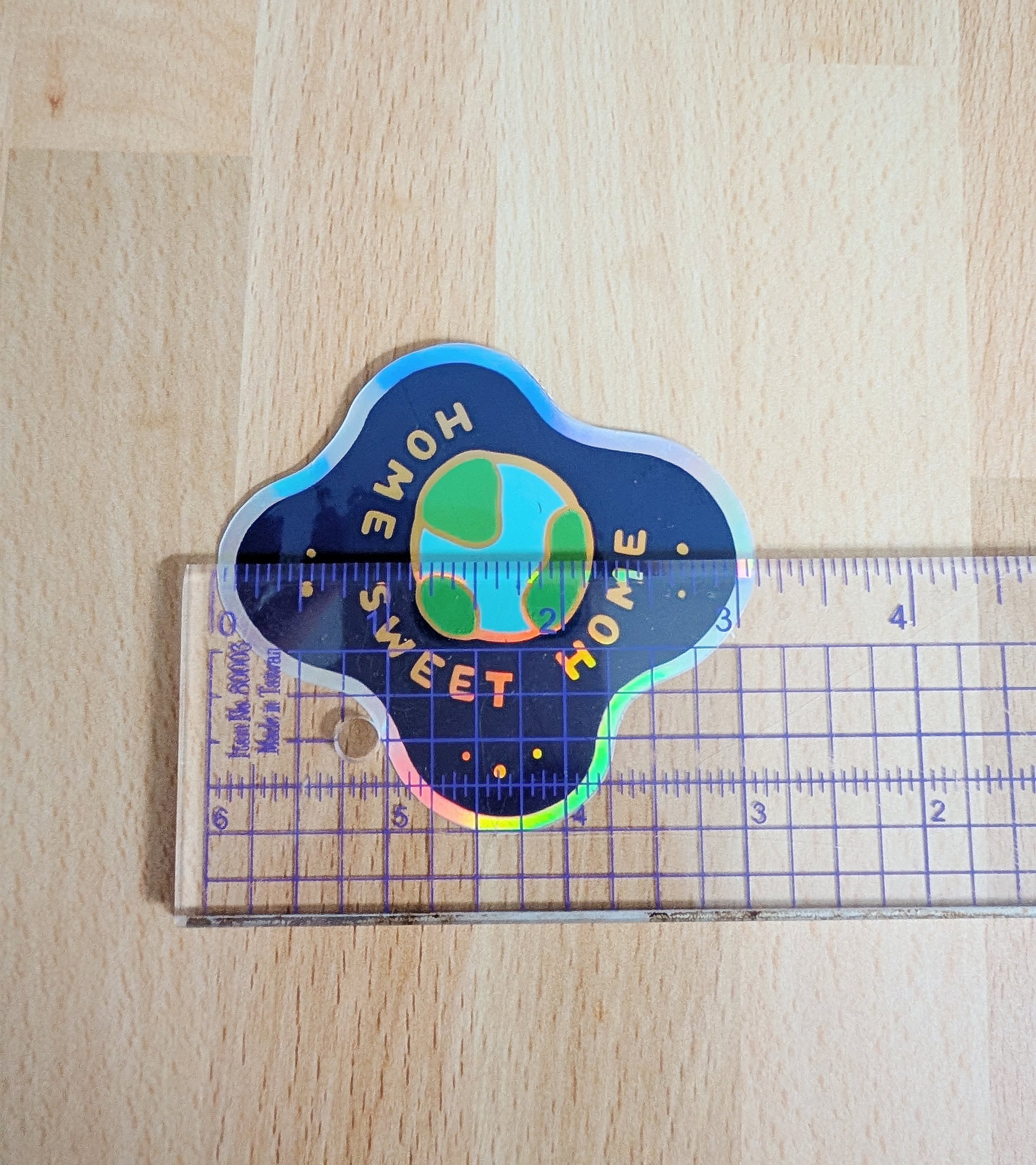 Holographic vinyl sticker featuring the planet earth floating in space with the words Home Sweet Home. There is a clear ruler on top of the sticker and the sticker measures 3 inches wide.