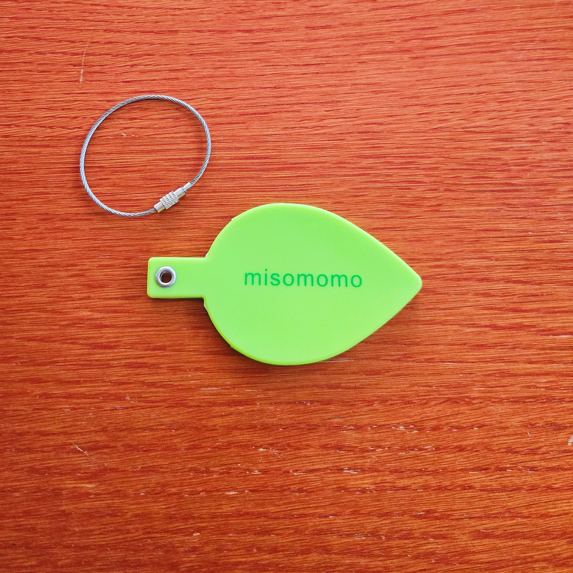 Green silicone leaf shaped luggage tag with the misomomo brand name/label. Luggage tag has a metal grommet that attaches to a sturdy metal keychain.