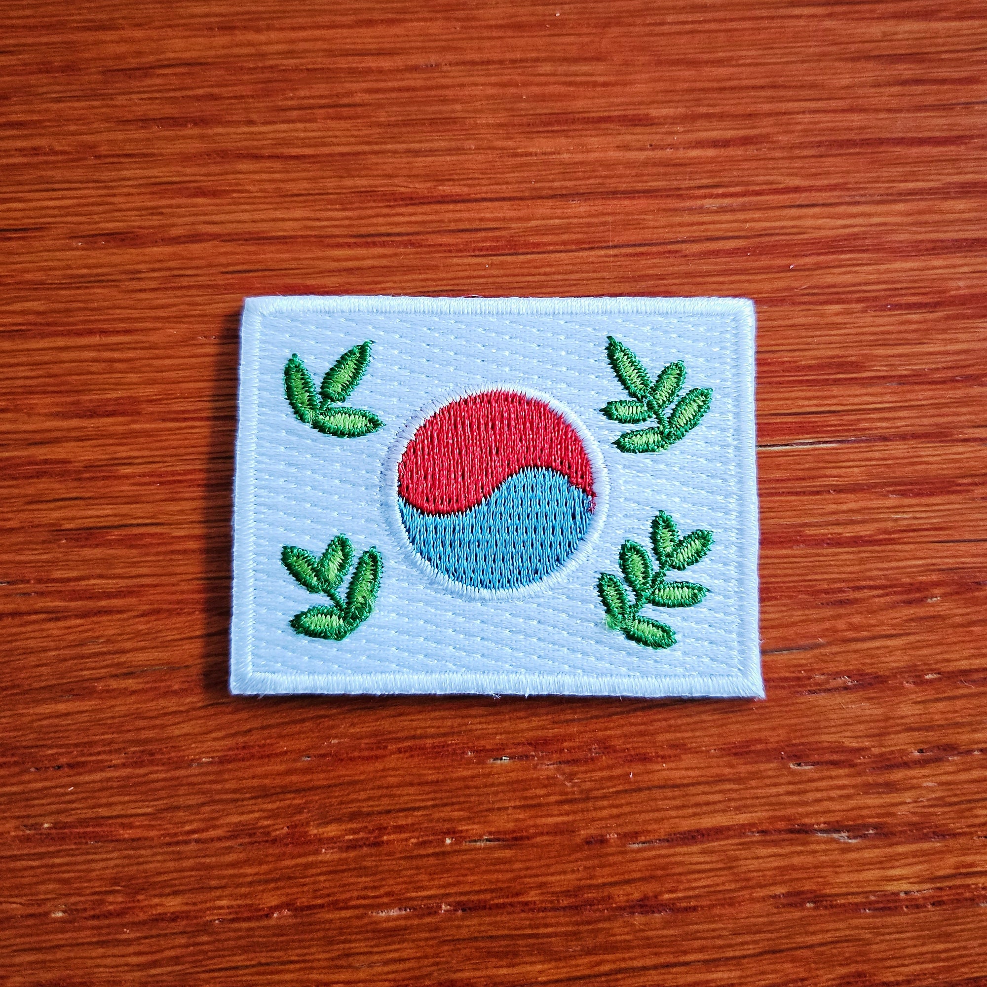 A small, iron-on patch featuring a stylized leafy design of the Korean flag with a green, red, and blue color scheme on a white background.