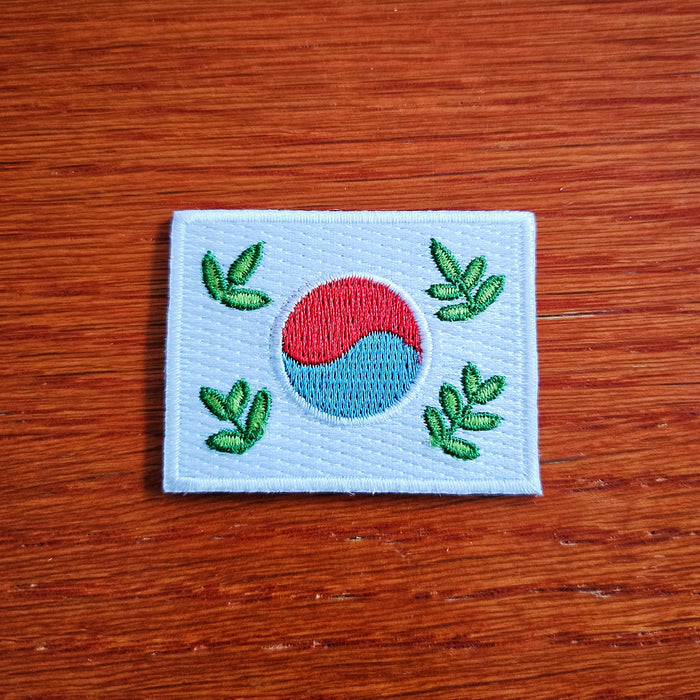A small, iron-on patch featuring a stylized leafy design of the Korean flag with a green, red, and blue color scheme on a white background.
