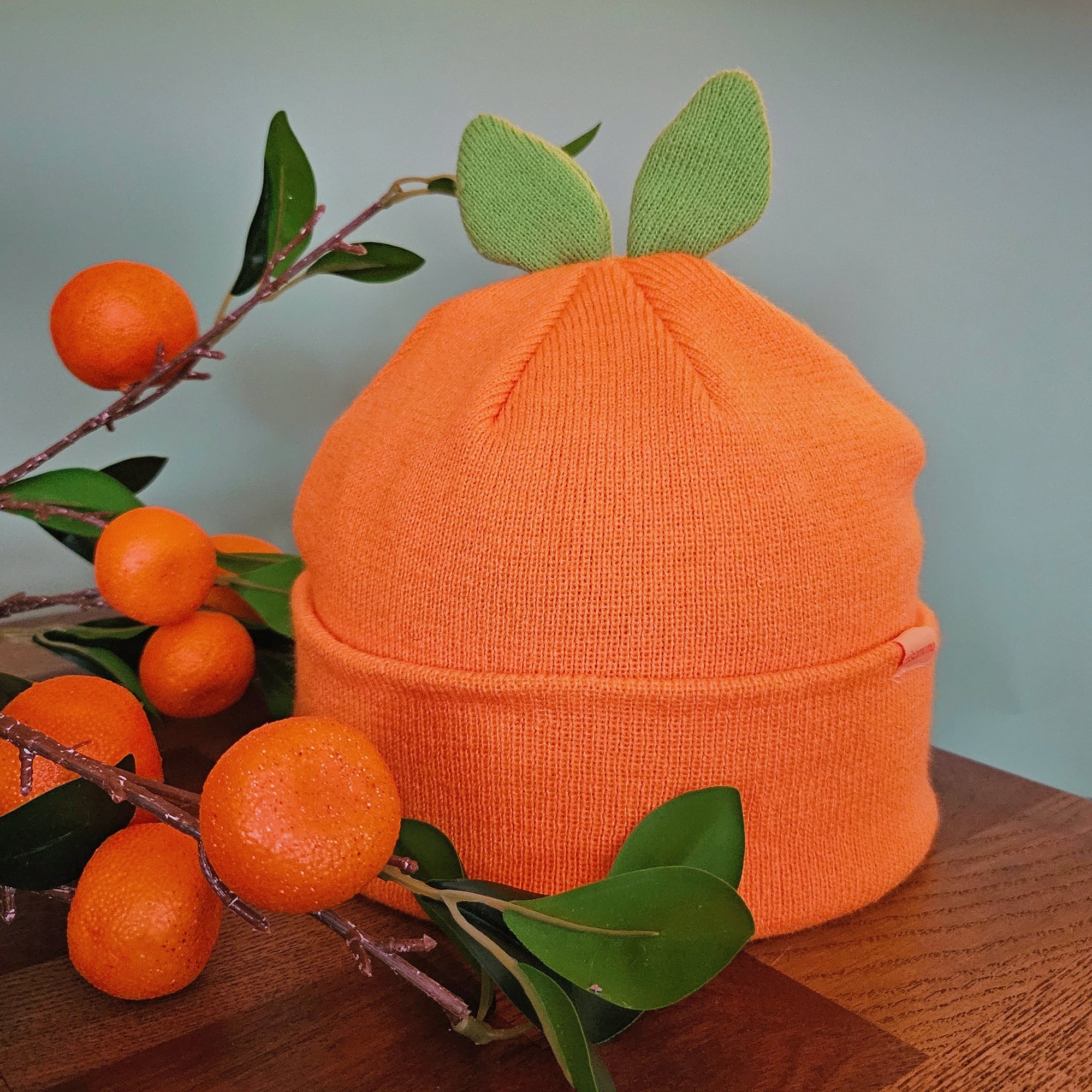 A cute orange beanie with two green leaves attached to the top part, so that it resembles an orange fruit when worn. Displayed on a wooden surface next to some orange or tangerine branches