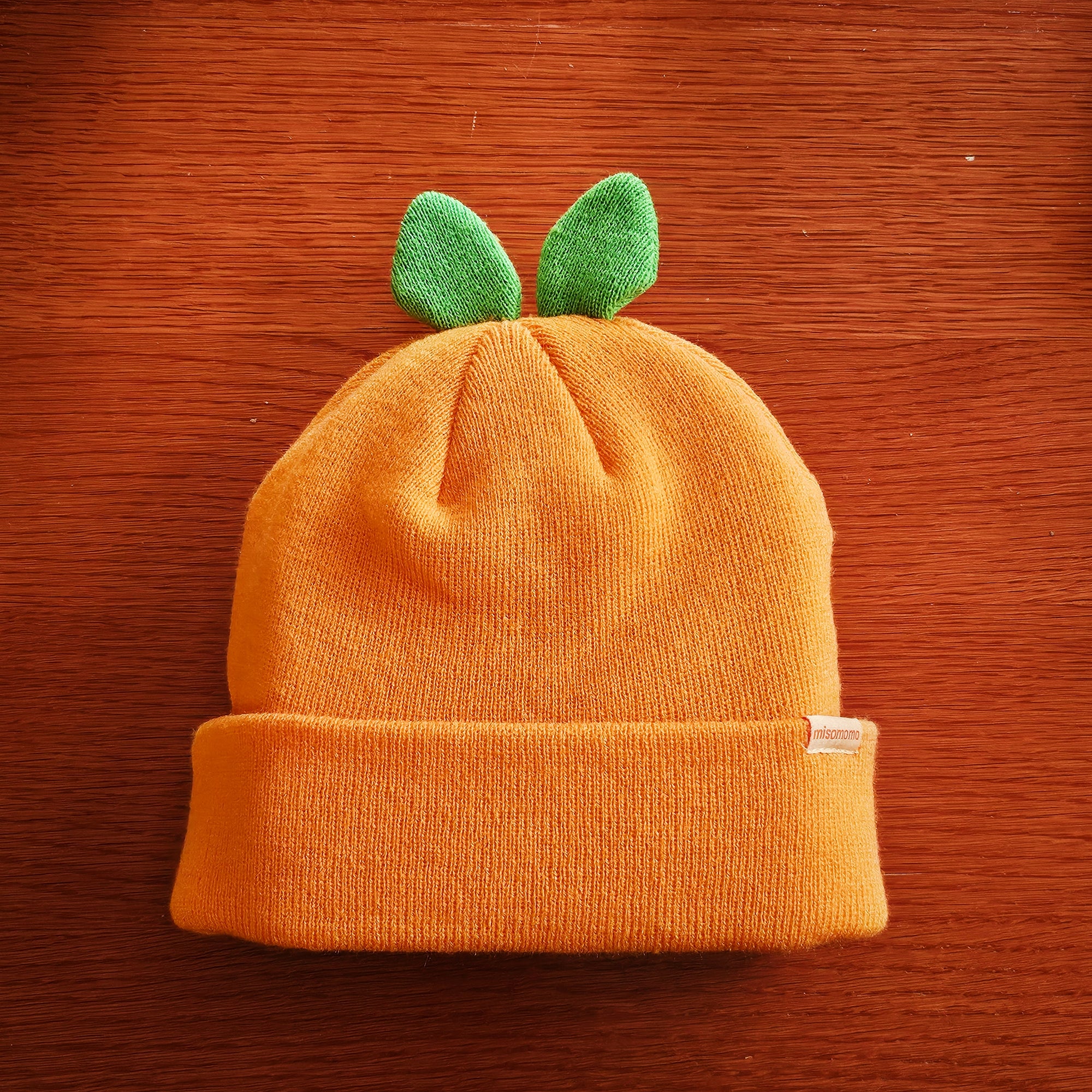 A cute orange beanie with two green leaves attached to the top part, so that it resembles an orange fruit when worn. Displayed on a wooden background.