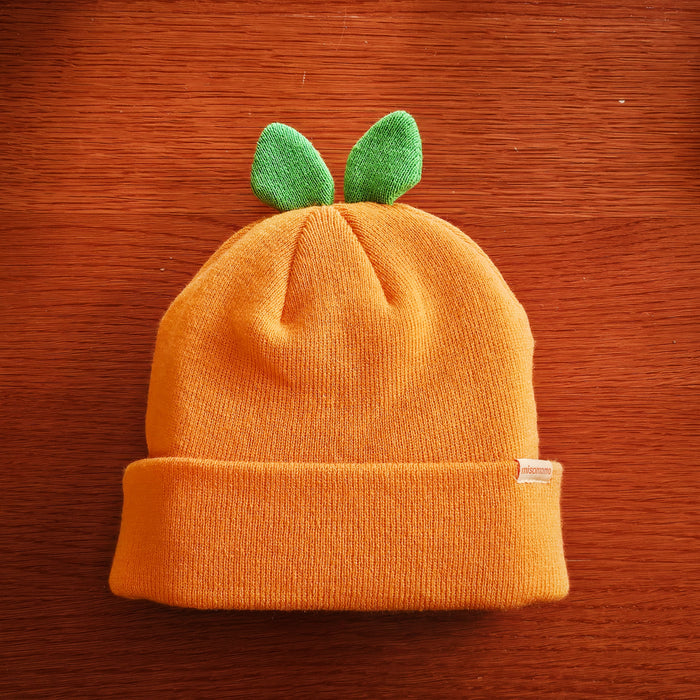 A cute orange beanie with two green leaves attached to the top part, so that it resembles an orange fruit when worn. Displayed on a wooden background.