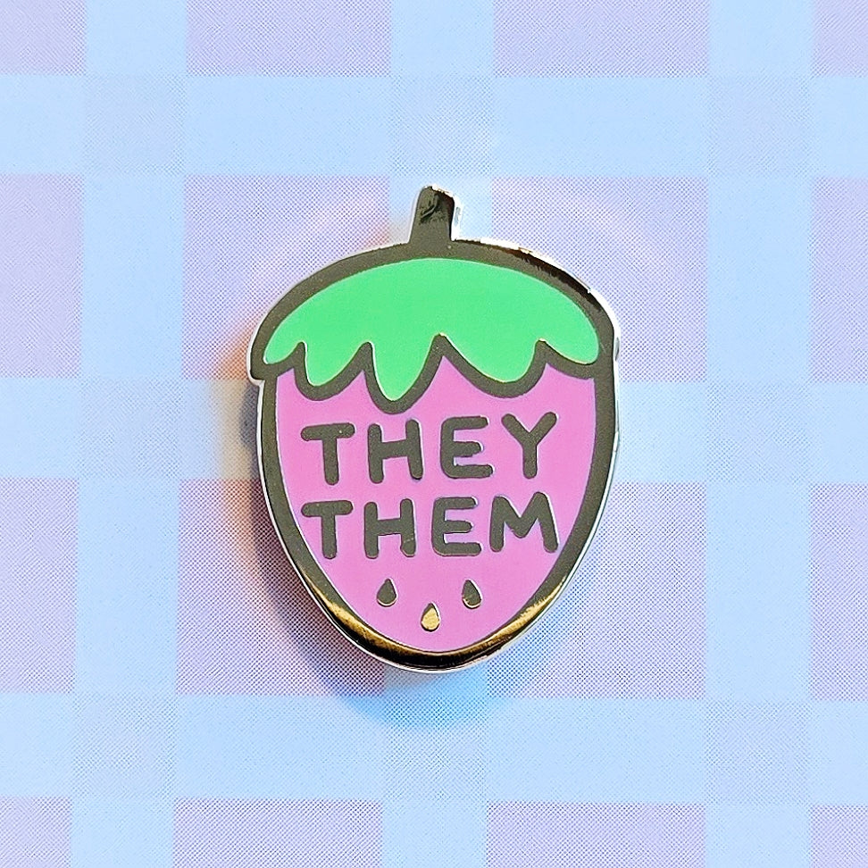 Enamel pin with gold plating, shaped like a cute strawberry in pink and green. Pronoun pin featuring they/them pronouns.