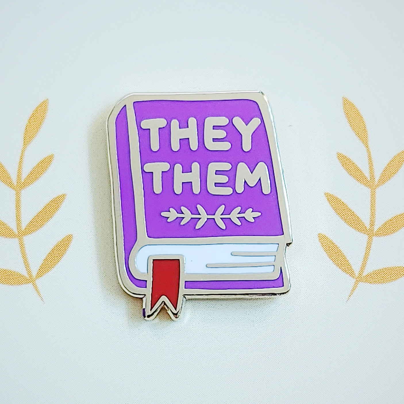 Purple misomomo pronoun enamel pin in the shape of a book with a red bookmark, featuring they/them pronouns