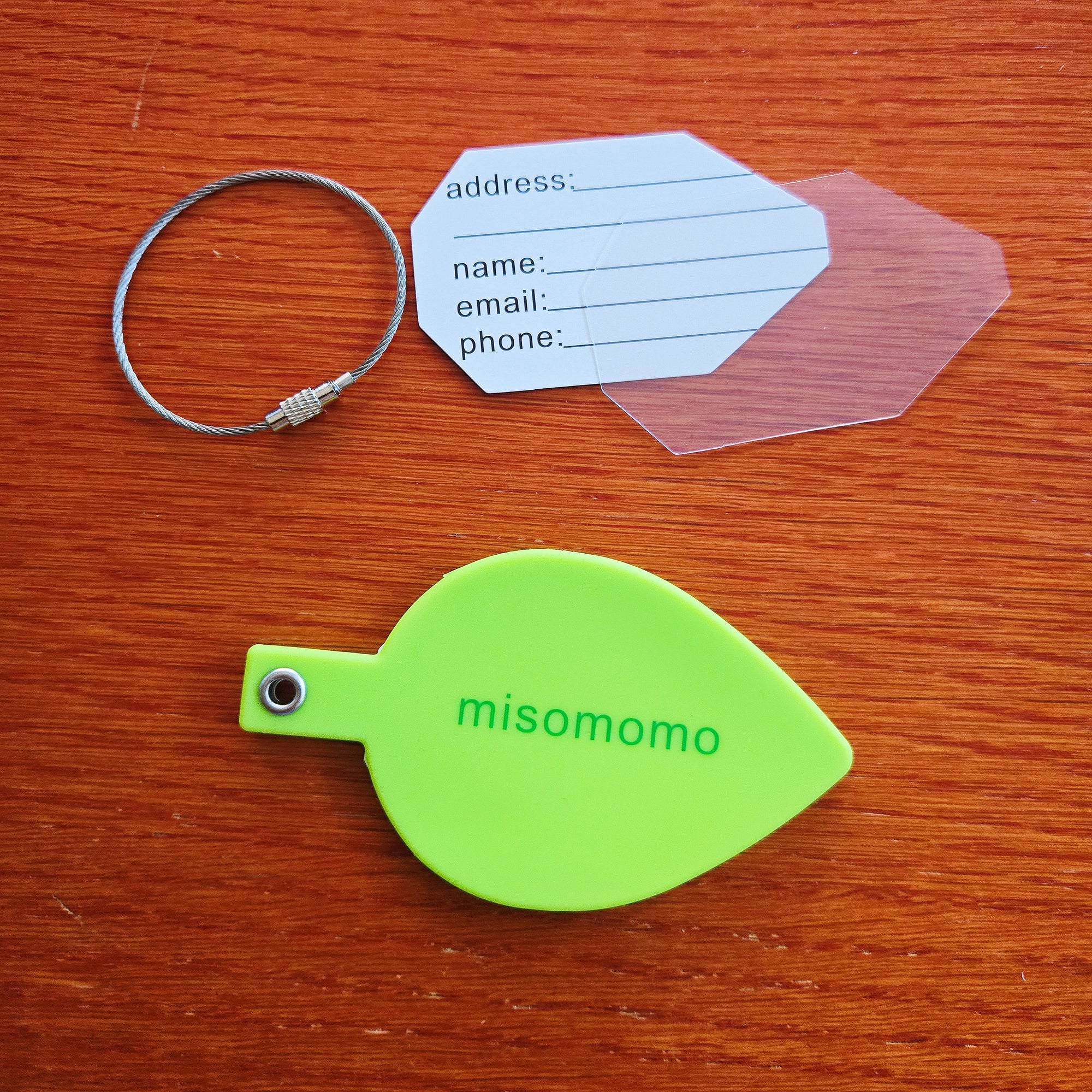 A green silicone leaf shaped luggage tag with the "misomomo" brand name/logo, and the included parts: a sturdy metal keychain, a paper tag for personal info, and a protective plastic cover for the written tag. The logo side of the luggage tag is showing.