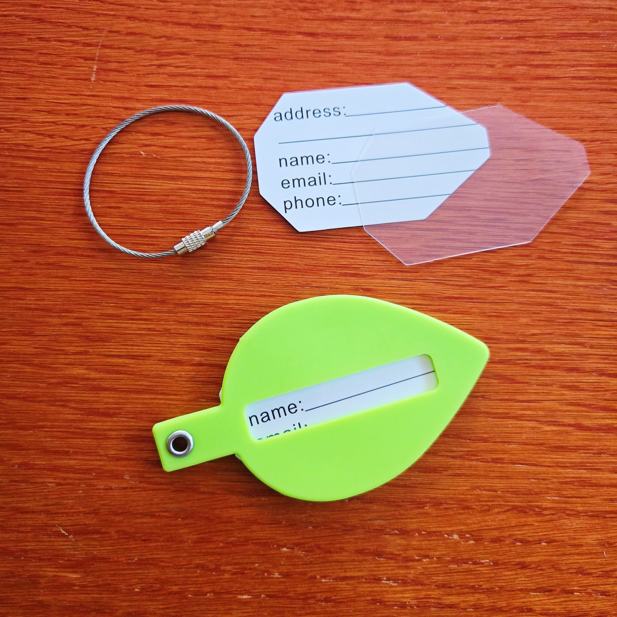 A green silicone leaf shaped luggage tag with the "misomomo" brand name/logo, and the included parts: a sturdy metal keychain, a paper tag for personal info, and a protective plastic cover for the written tag. The name tag cutout side of the luggage tag is showing.