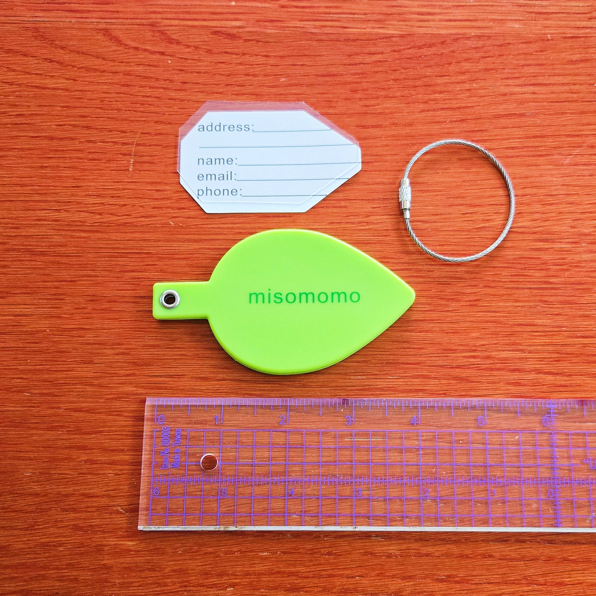 A green silicone leaf shaped luggage tag with the "misomomo" brand name/logo, and the included parts: a sturdy metal keychain, a paper tag for personal info, and a protective plastic cover for the written tag. There is a clear ruler and the leaf measures four inches long