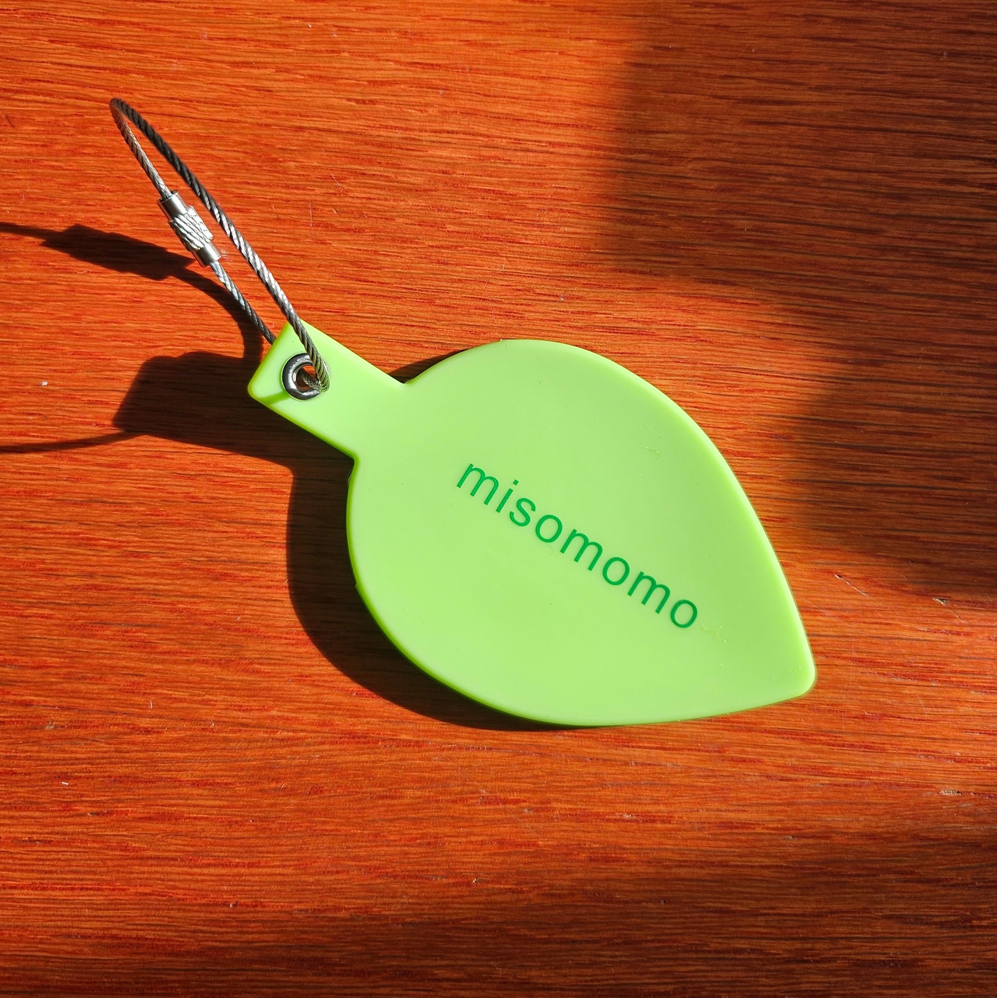 A green leaf-shaped luggage tag with the 'misomomo' brand name on it, featuring a metal grommet and attached to a stainless steel cable, placed on a wooden surface.
