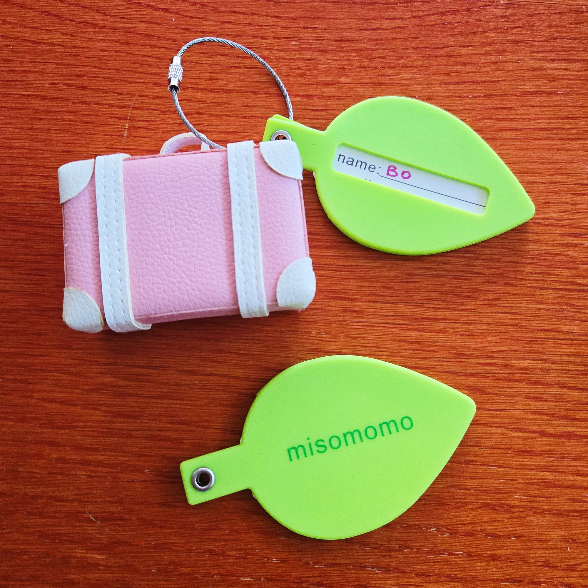 Green silicone leaf-shaped luggage tags with the misomomo logo on one side and a tag for your personal info. Pictured attached to a tiny suitcase.