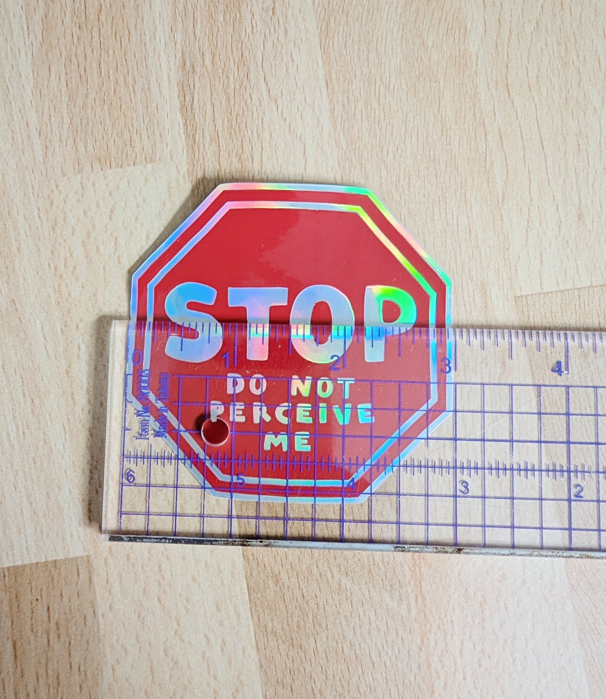 holographic red vinyl sticker in the shape of a red stop sign, with the phrase "STOP do not perceive me". There is a clear ruler on top of the sticker and the sticker measures 3 inches wide