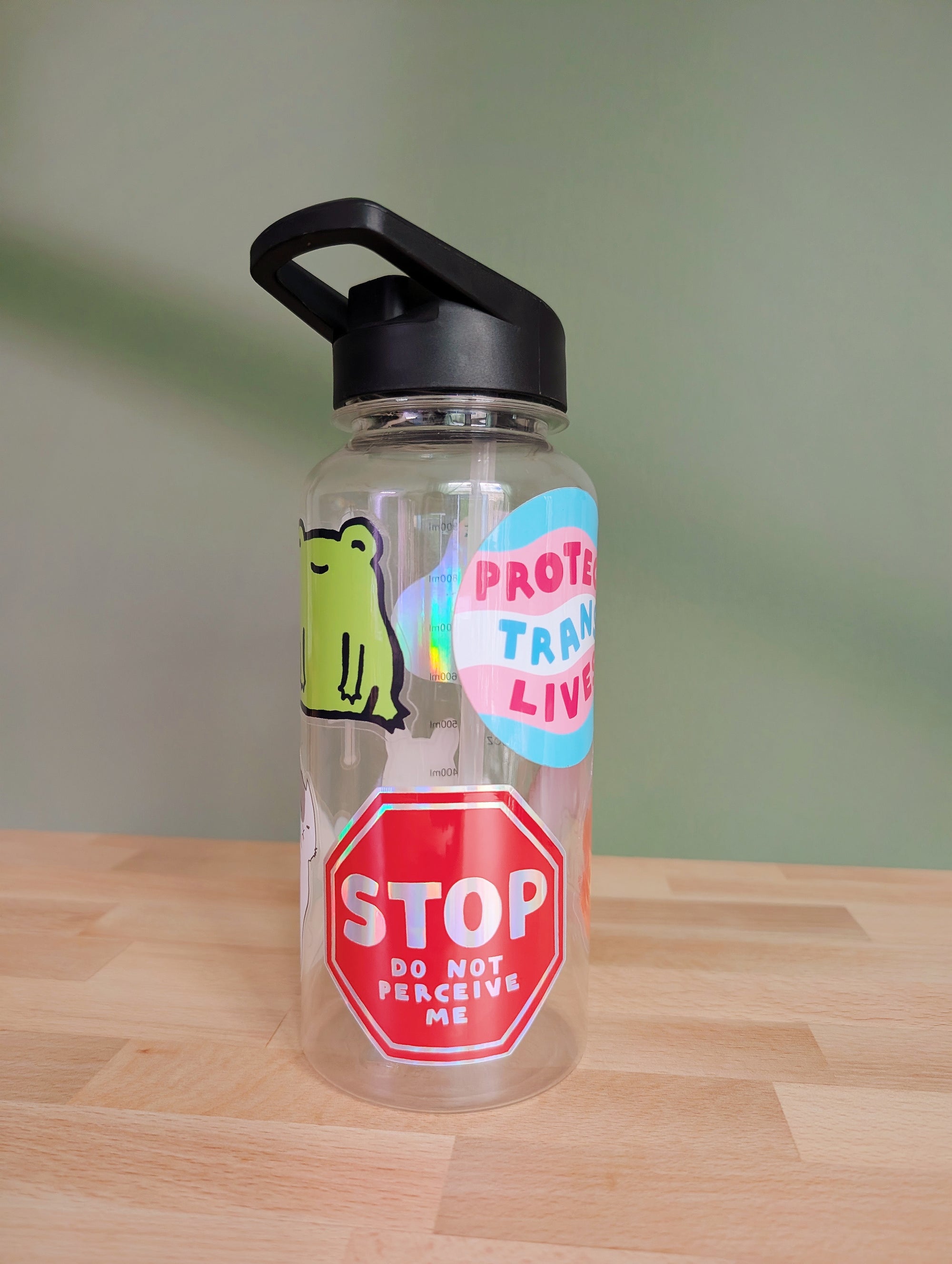 a clear water bottle with a bunch of vinyl stickers on it as decoration. on the front is a red holographic sticker in the shape of a stop sign that says "STOP do not perceive me"