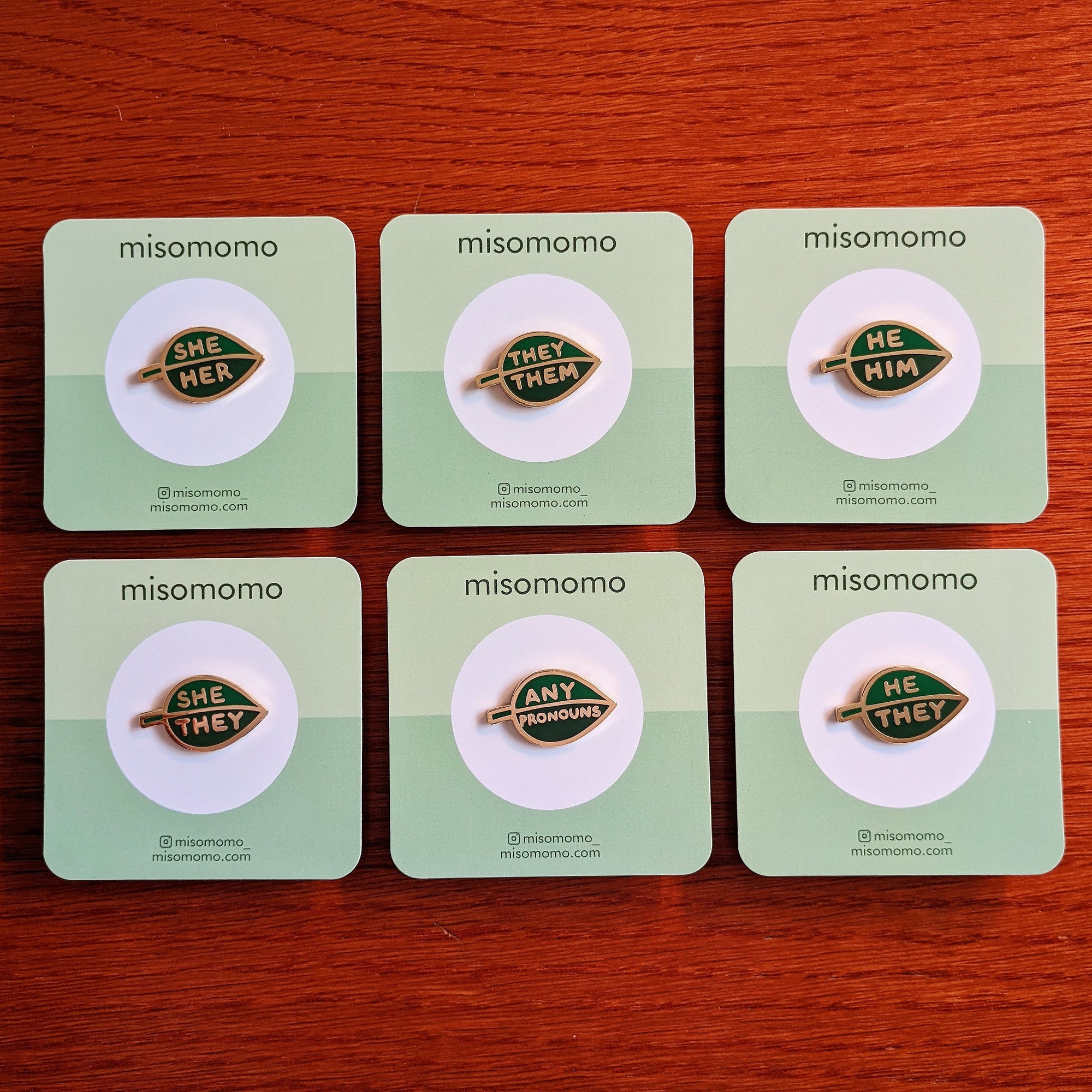 group of six different misomomo brand leaf shaped enamel pride pins, with different pronoun combinations. all pins are on backing cards and laid out on a wooden surface.