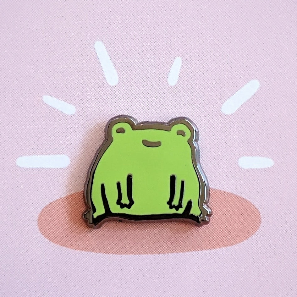 a cute misomomo enamel pin in the shape of a happy little green frog, sitting on the ground and smiling
