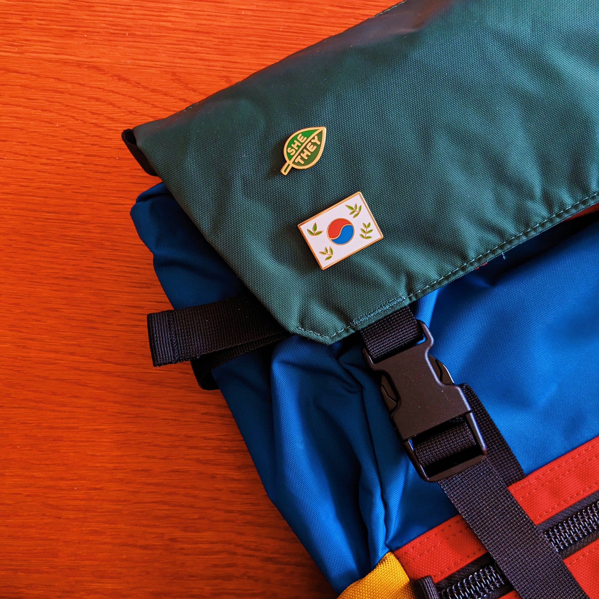 a colorful green/blue/red backpack featuring two misomomo enamel pins, a leafy green korean flag enamel pin and a green leaf-shaped she/they pronoun pin