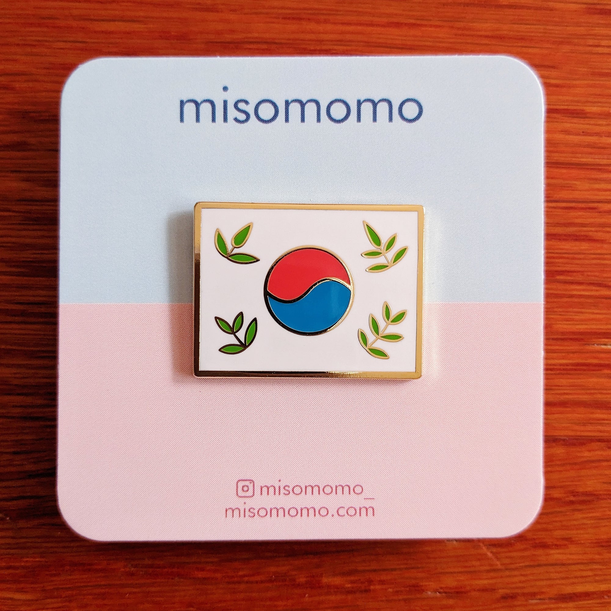 Stylized leafy Korean flag (taegukgi) enamel pin pictured on the misomomo backing card on a blue and pink background