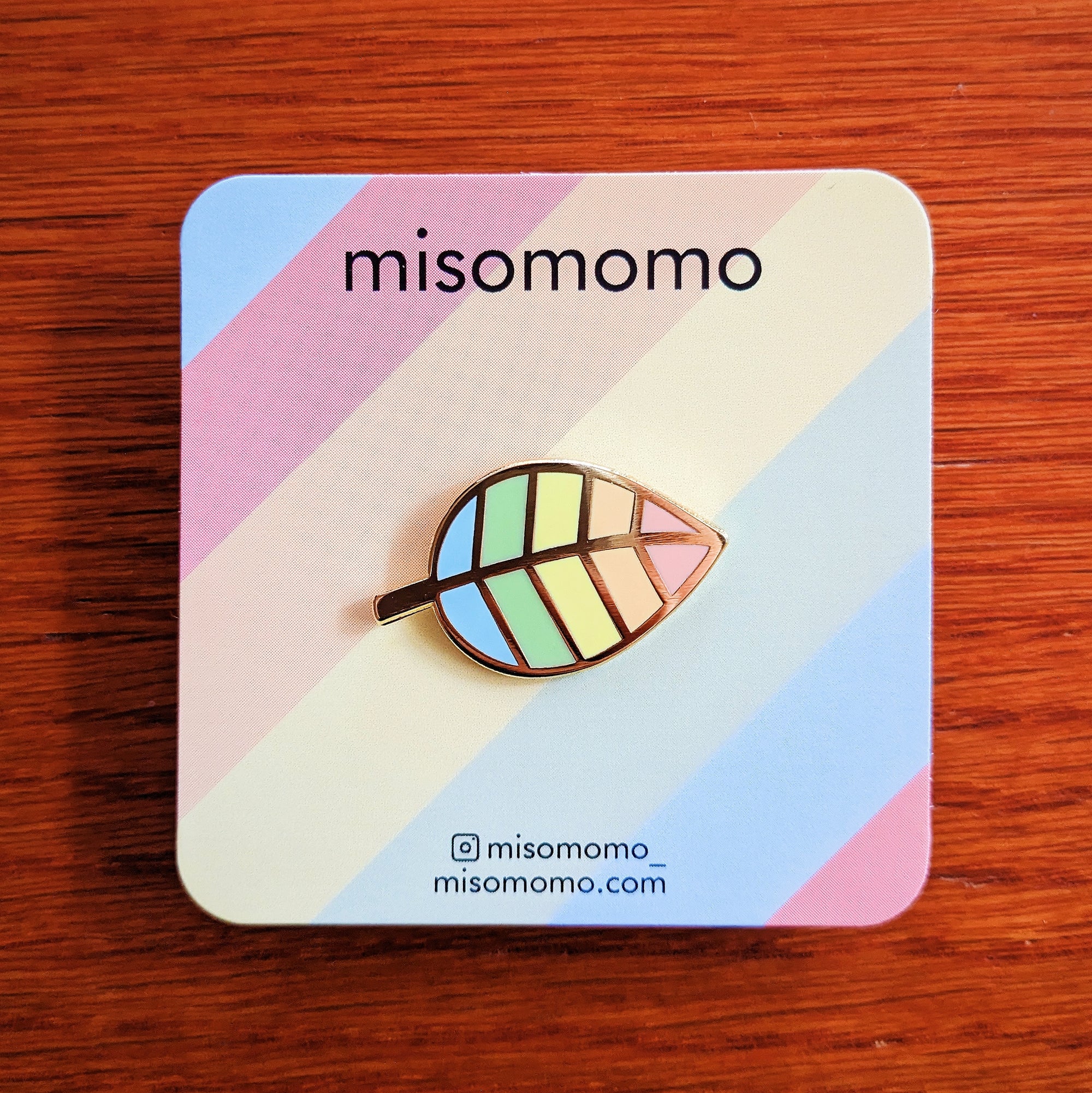 misomomo enamel pin in the shape of a leaf, with pastel rainbow colors, pictured on the rainbow backing card